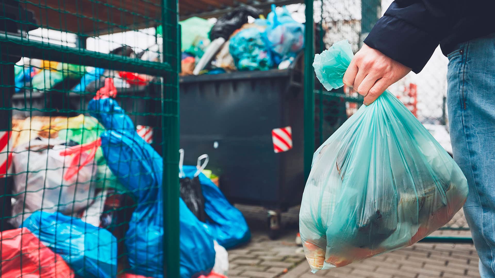 Una persona con in mano un sacchetto celeste di spazzatura si avvicina ad una zona di scarico per buttare via i rifiuti.